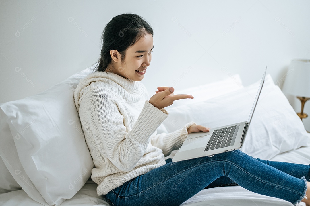 Mulher vestindo uma camisa branca na cama e jogando laptop alegremente.