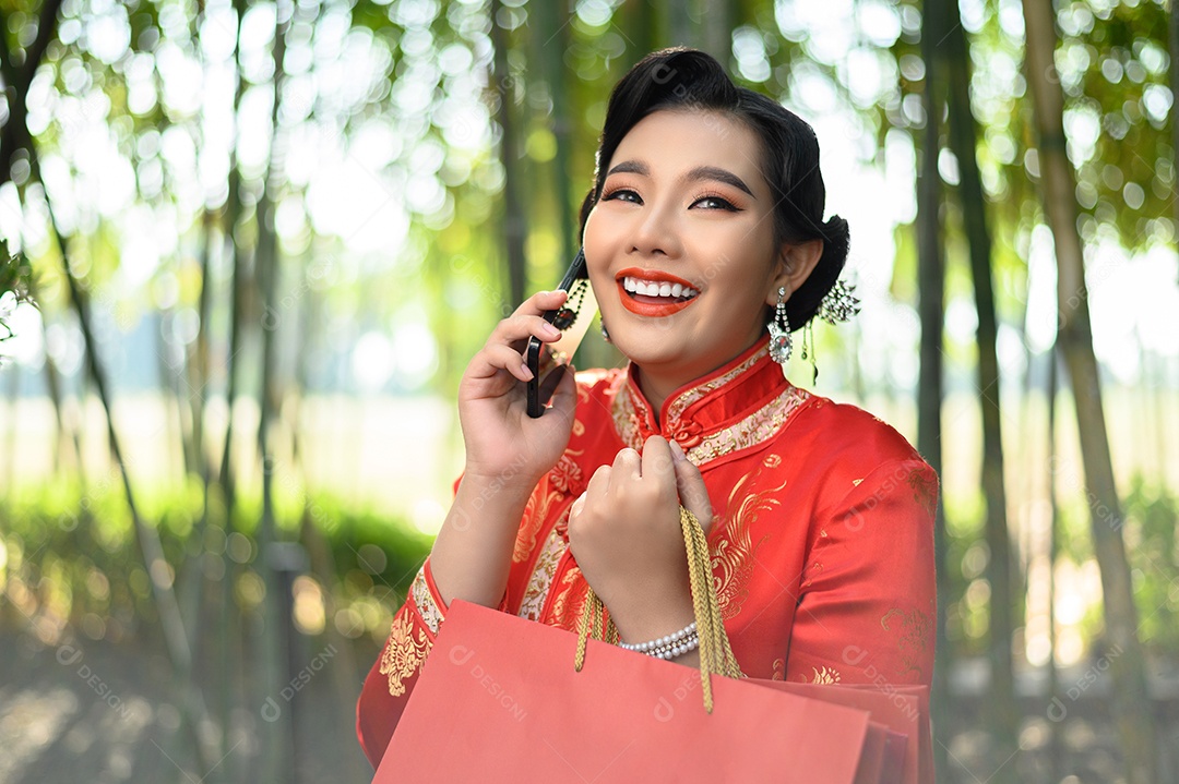 Mulher asiática bonita em um cheongsam chinês segurando sacolas de compras e falando no celular com felicidade no fundo da floresta