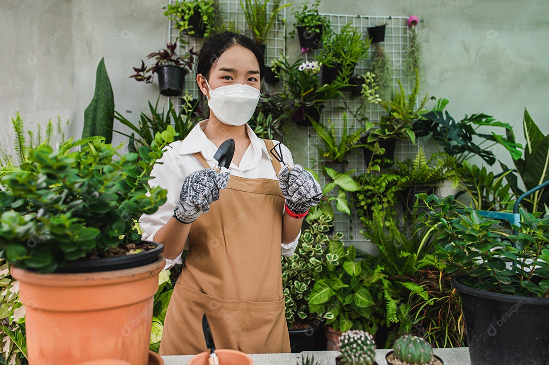 Mulher jardineira asiática usando máscara facial e suporte de avental e mostrando uma pá e forcado, equipamento para transplantes de plantas de interior