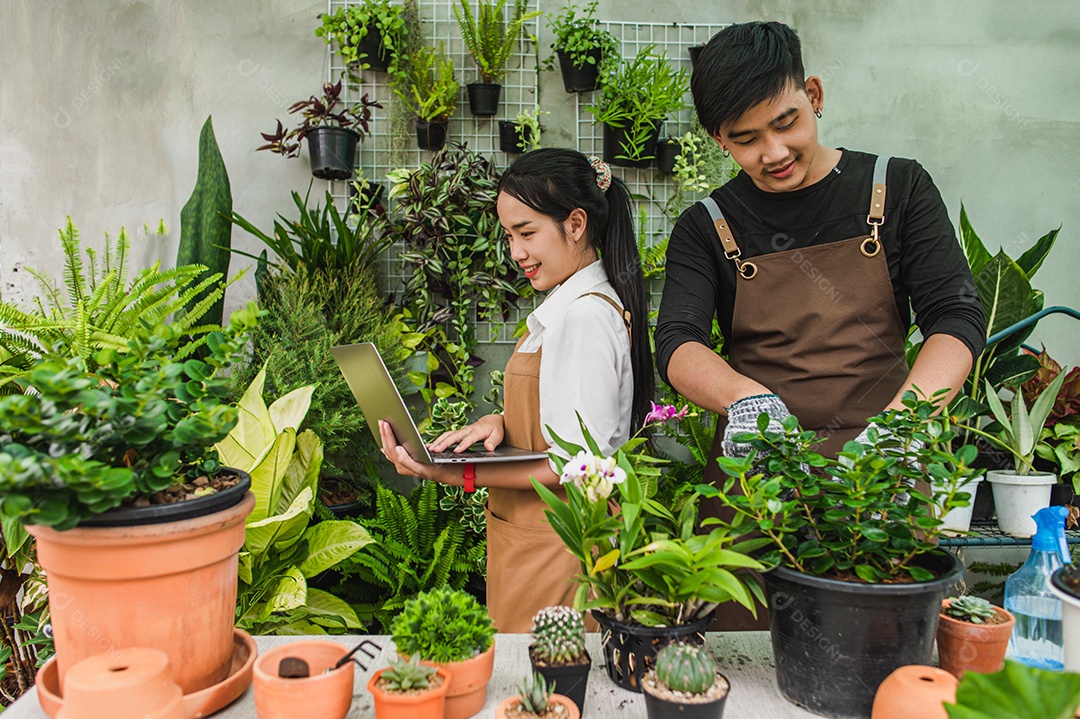 Feliz casal de jovens jardineiros asiáticos usando avental usa equipamento de jardim e computador portátil para cuidar das plantas da casa na loja