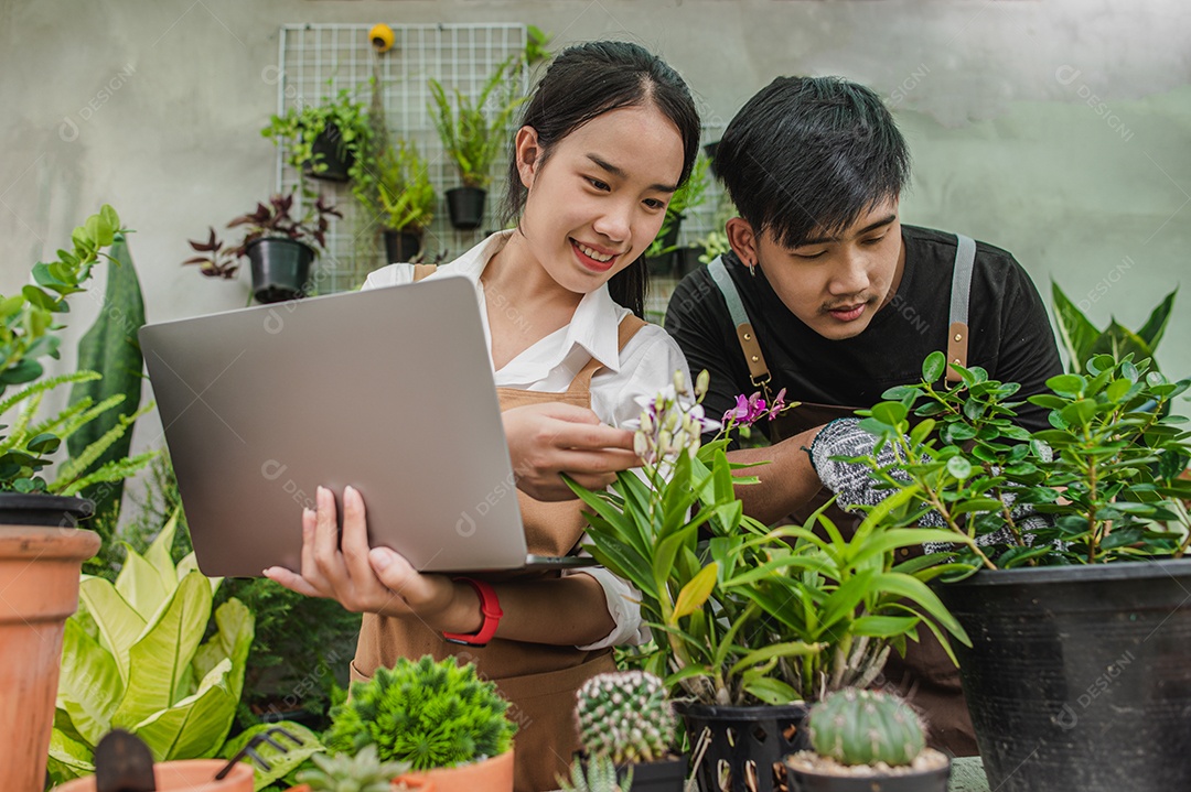 Feliz casal de jovens jardineiros asiáticos usando avental usa equipamento de jardim e computador portátil para cuidar das plantas da casa na loja