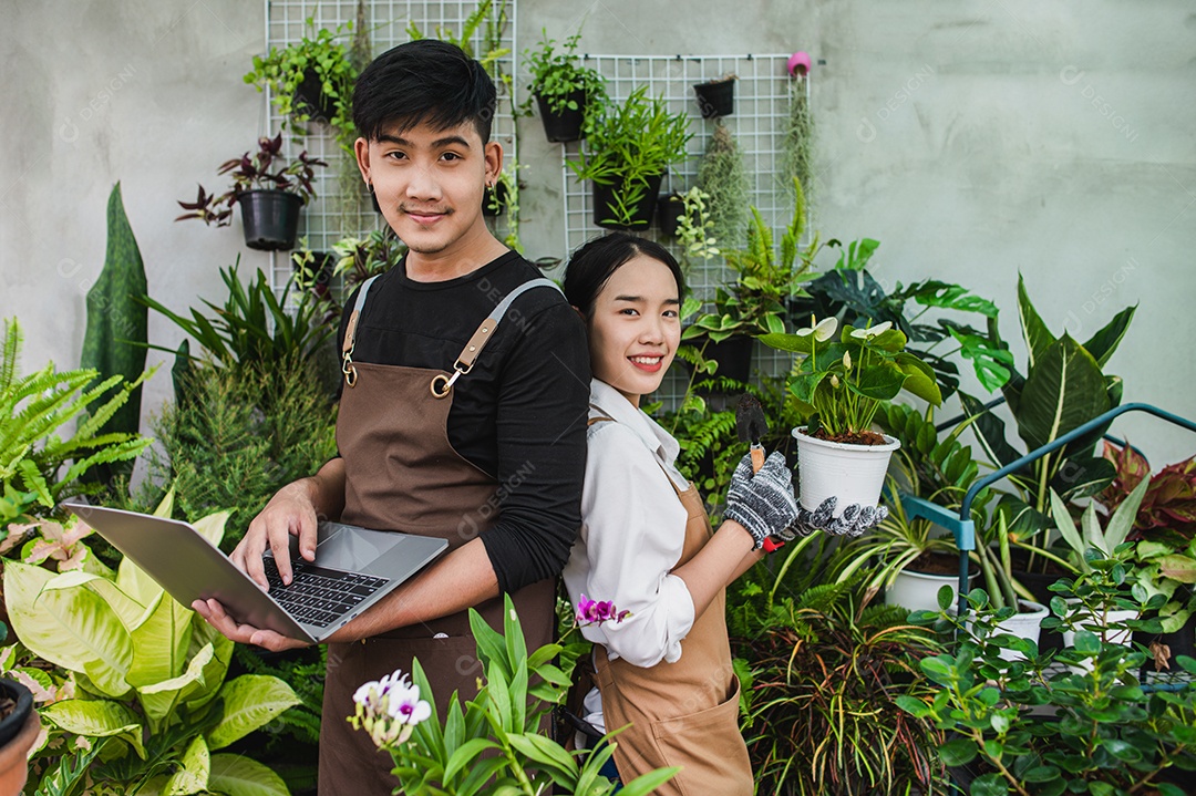 Retrato casal de jovens jardineiros asiáticos usando avental usa equipamento de jardim e computador portátil para pesquisar e cuidar das plantas