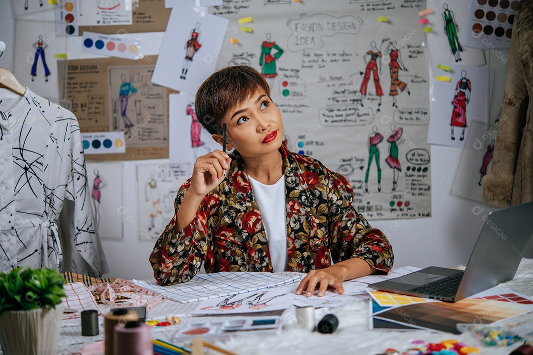 Jovem designer feminina usa lápis de toque no templo durante o trabalho com o computador portátil para procurar ideias para roupas de design na mesa