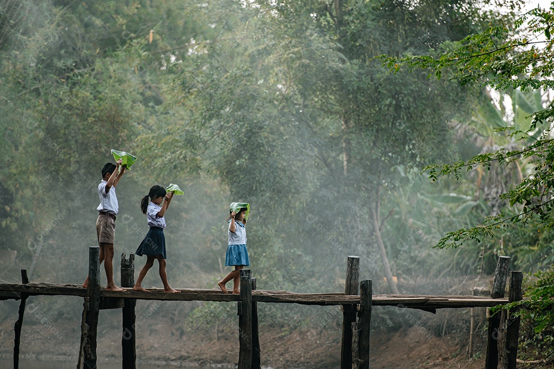 Menino asiático e duas meninas em uniforme de estudante e descalços segurando folha de bananeira na mão acima da cabeça, andando na ponte de madeira