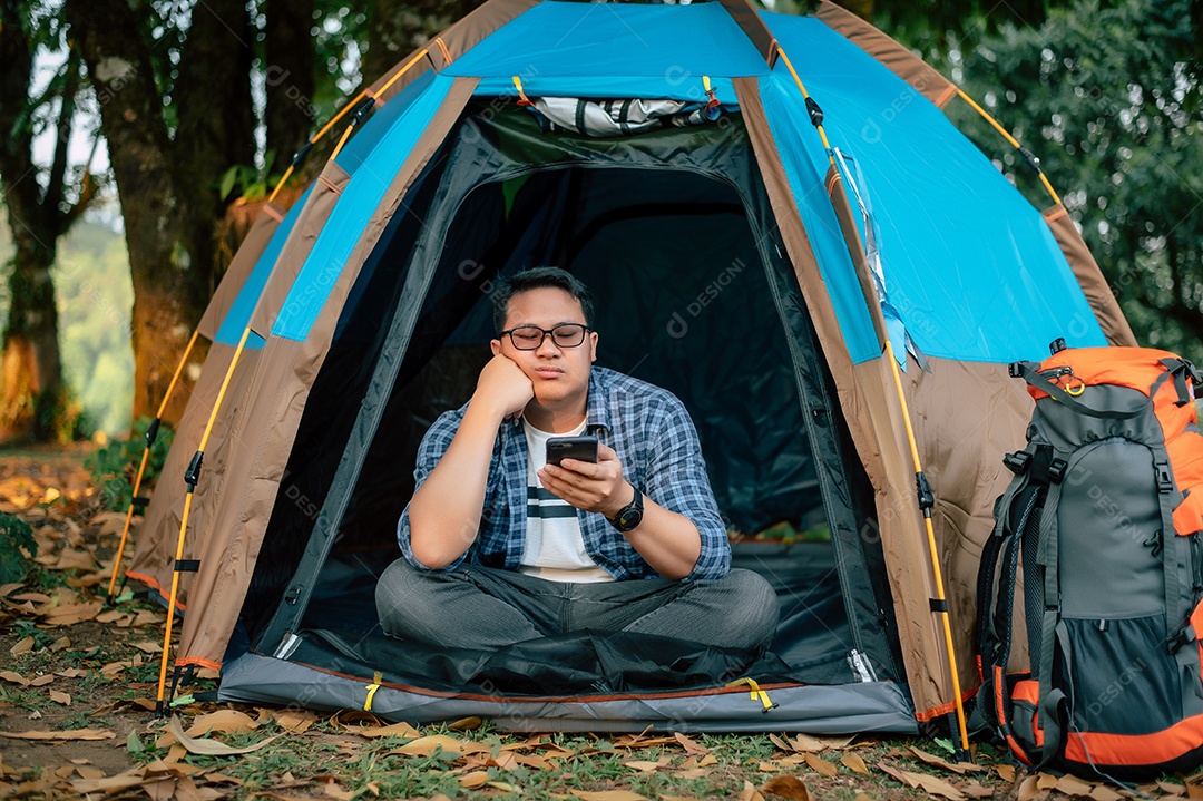 Retrato de óculos de homem viajante asiático entediado usando smartphone em acampamento de tenda. Conceito de viagens