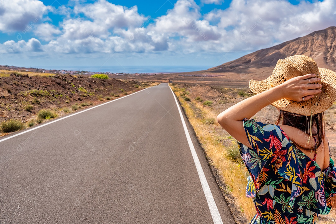 Vista traseira do desejo de viajar de garota hipster andando na estrada de asfalto em terras selvagens na ilha de Fuerteventura - turista feminina com chapéu viajando nas Ilhas Canárias pedindo carona no caminho - conceito de viagem