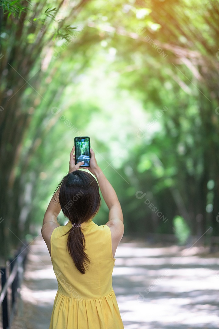 Mulher asiática de vestido amarelo e chapéu viajando no túnel de bambu verde, viajante feliz tirando foto pelo celular no templo, Tailândia