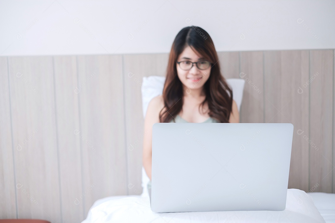 Feliz mulher asiática sentada e usando computador laptop na cama branca