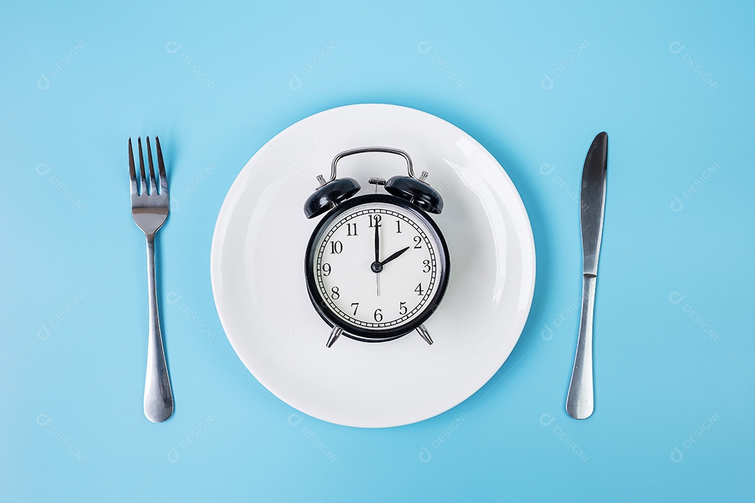 Vista superior despertador na chapa branca com garfo e faca sobre fundo azul. Jejum intermitente, dieta cetogênica, perda de peso