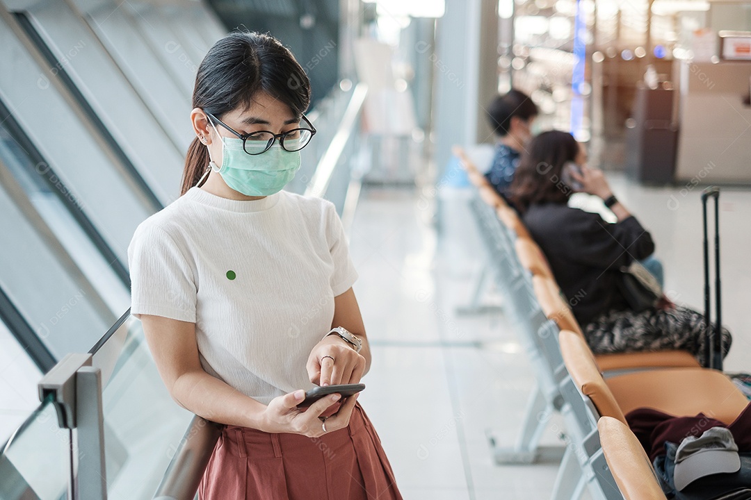 Jovem mulher usando máscara facial e usando smartphone móvel no aeroporto, proteção contra infecção por doença de coronavírus (Covid-19). Novo normal e distanciamento social