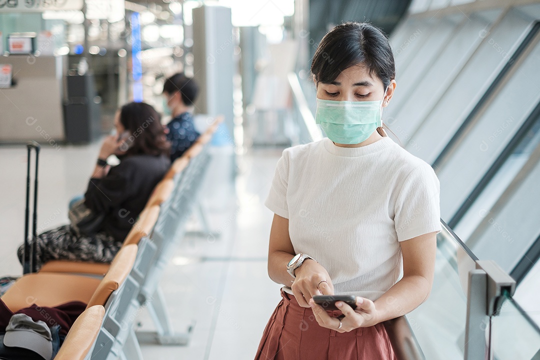 Jovem mulher usando máscara facial e usando smartphone móvel no aeroporto, proteção contra infecção por doença de coronavírus (Covid-19). Novo normal e distanciamento social