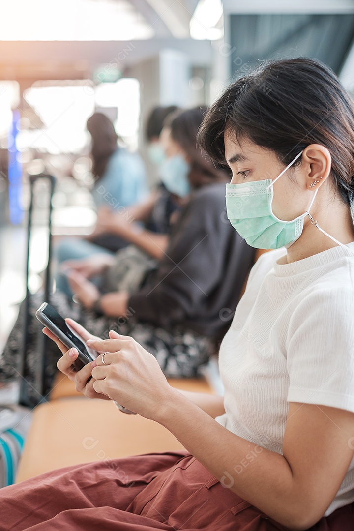 Jovem mulher usando máscara facial e usando smartphone móvel no aeroporto, proteção contra infecção por doença de coronavírus (Covid-19). Novo normal e distanciamento social