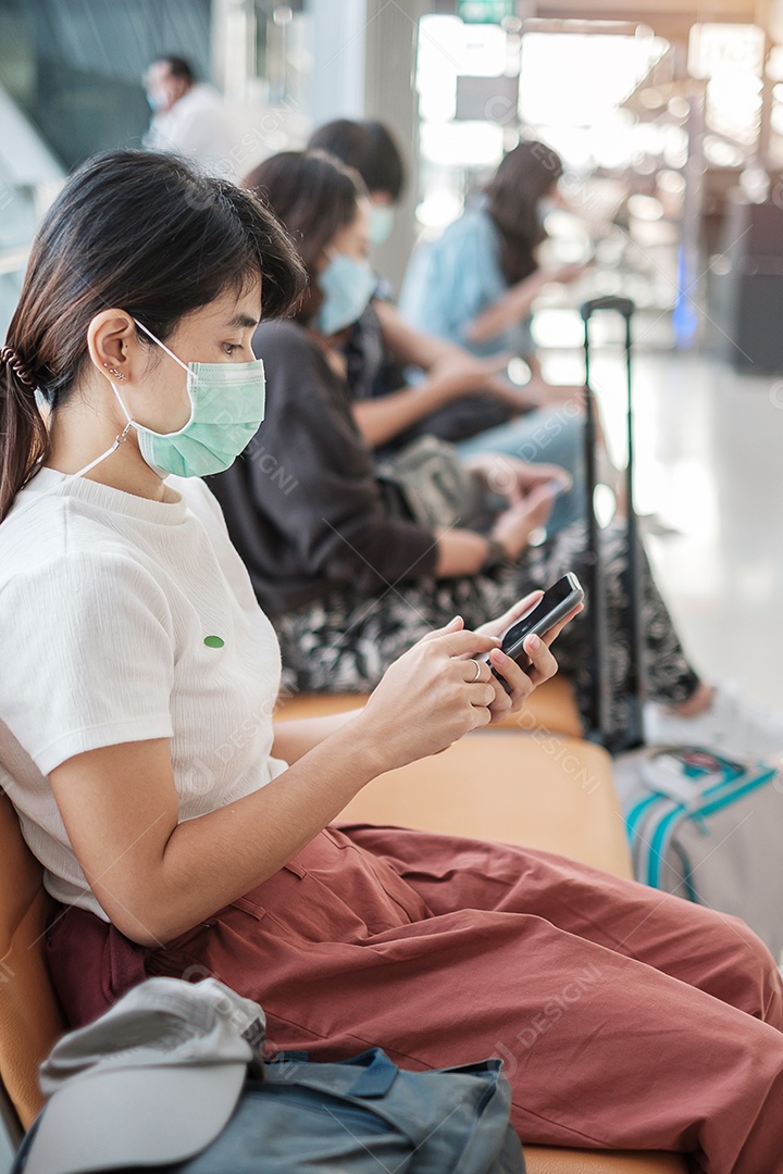 Jovem mulher usando máscara facial e usando smartphone móvel no aeroporto, proteção contra infecção por doença de coronavírus (Covid-19). Novo normal e distanciamento social