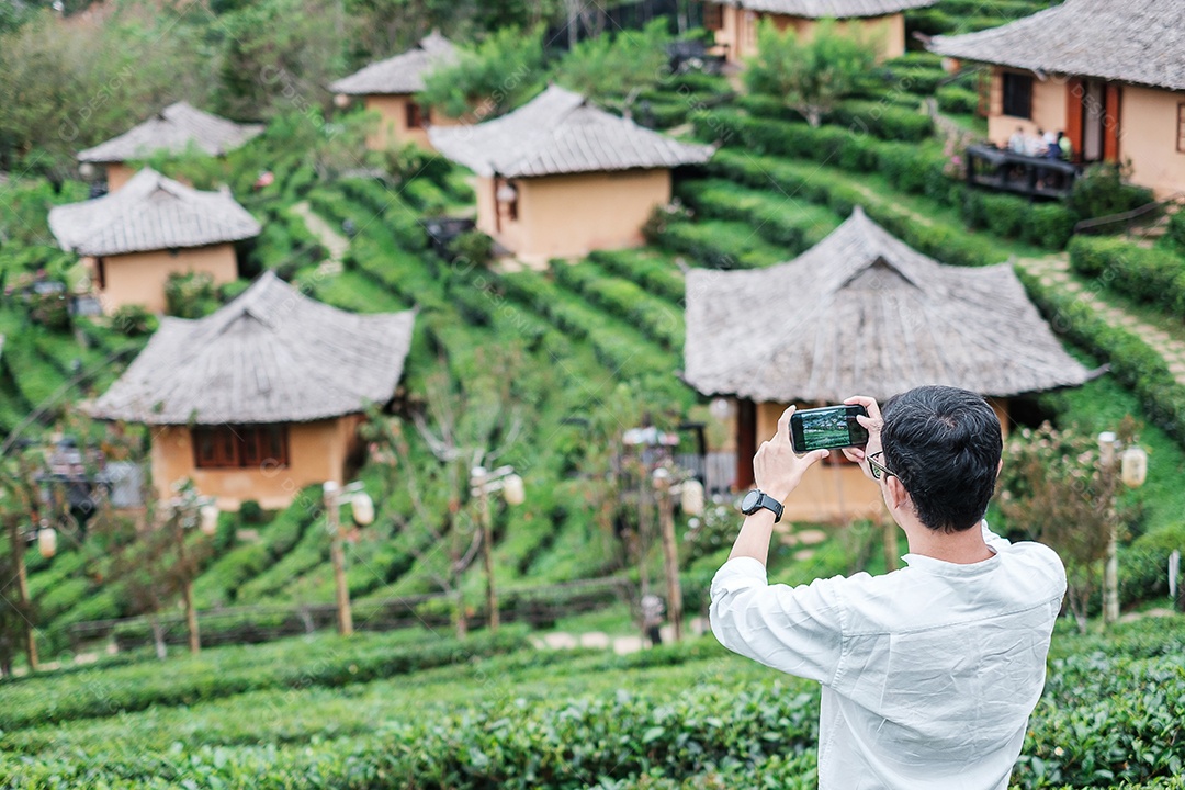 Homem de turista feliz na camisa branca tirando foto pelo smartphone móvel no belo jardim de chá. Viajante visitando a vila tailandesa