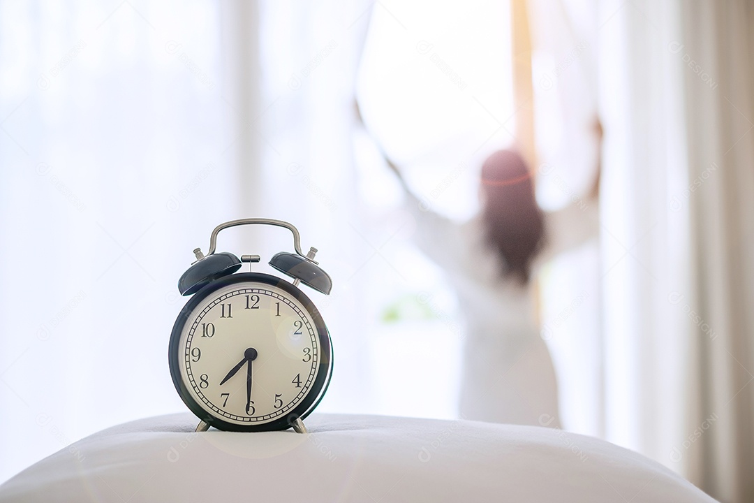 Closeup despertador com mulher feliz, estendendo-se no quarto depois de acordar, jovem adulto feminino levantando os braços e olhando para a janela de manhã. fresco relaxe e tenha um bom dia conceitos