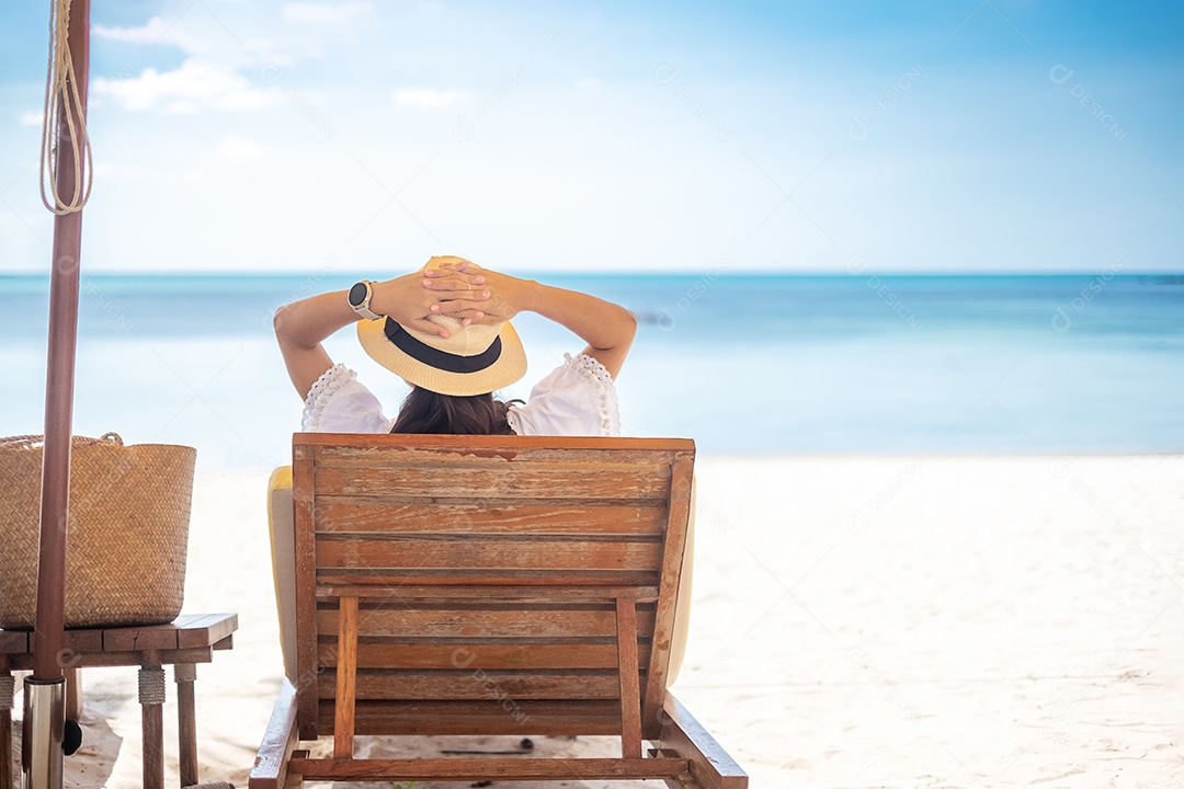 A mulher viajante feliz de vestido branco e chapéu desfruta de uma bela vista para o mar, jovem sentada na cadeira e olhando o oceano na praia tropical. Liberdade, relaxamento, férias de férias e conceito de viagens de verão