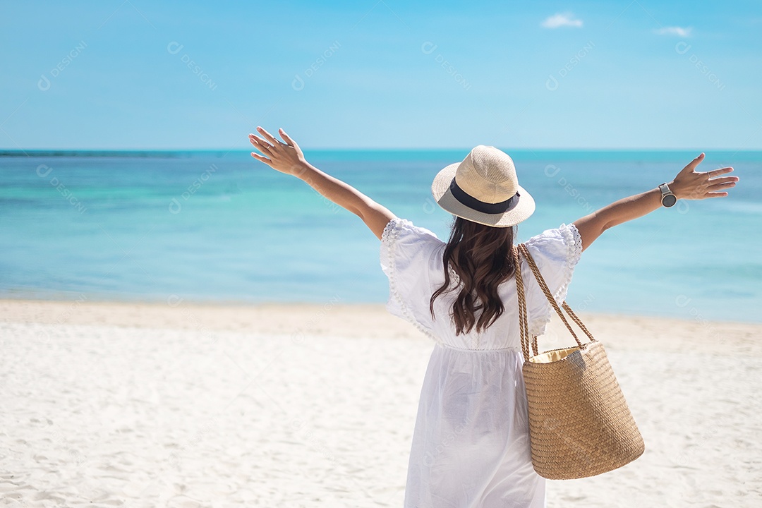 Feliz viajante mulher de vestido branco e chapéu desfrutar de uma bela vista para o mar, jovem de pé na areia e olhando o oceano na praia tropical. Liberdade, relaxamento, férias de férias e conceito de viagens de verão