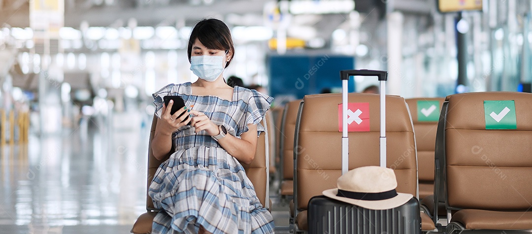 Jovem mulher usando máscara facial e usando smartphone móvel no aeroporto, proteção contra infecção por doença de coronavírus (Covid-19), viajante asiática sentada na cadeira. Novo normal e distanciamento social