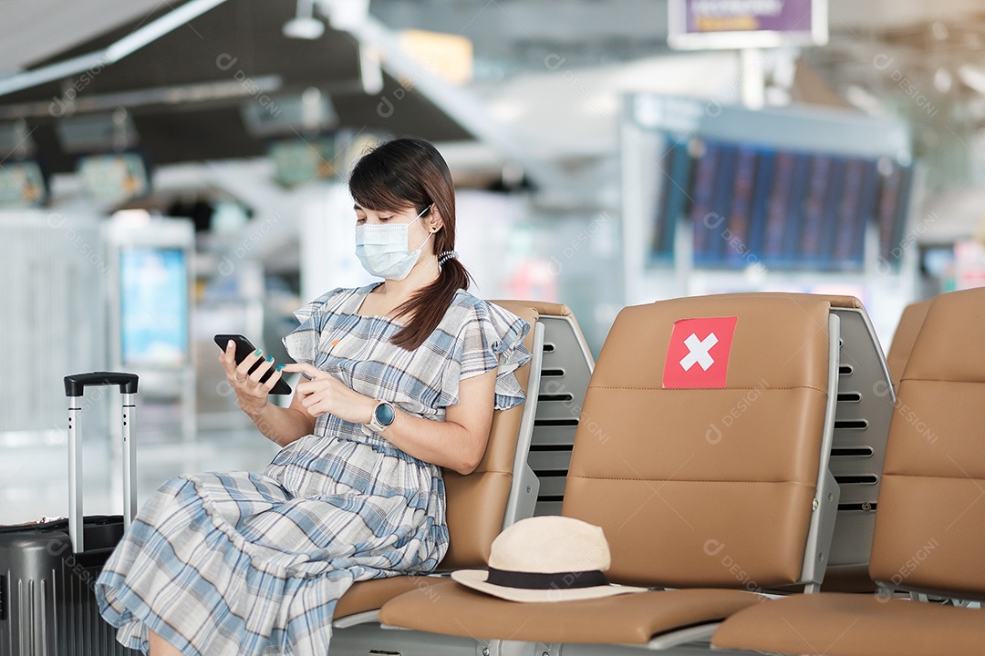 Jovem mulher usando máscara facial e usando smartphone móvel no aeroporto, proteção contra infecção por doença de coronavírus (Covid-19), viajante asiática sentada na cadeira. Novo normal e distanciamento social