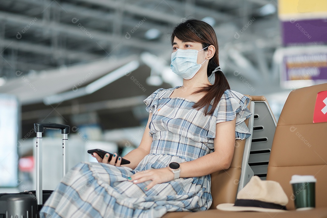 Jovem mulher usando máscara facial e usando smartphone móvel no aeroporto, proteção contra infecção por doença de coronavírus (Covid-19), viajante asiática sentada na cadeira. Novo normal e distanciamento social
