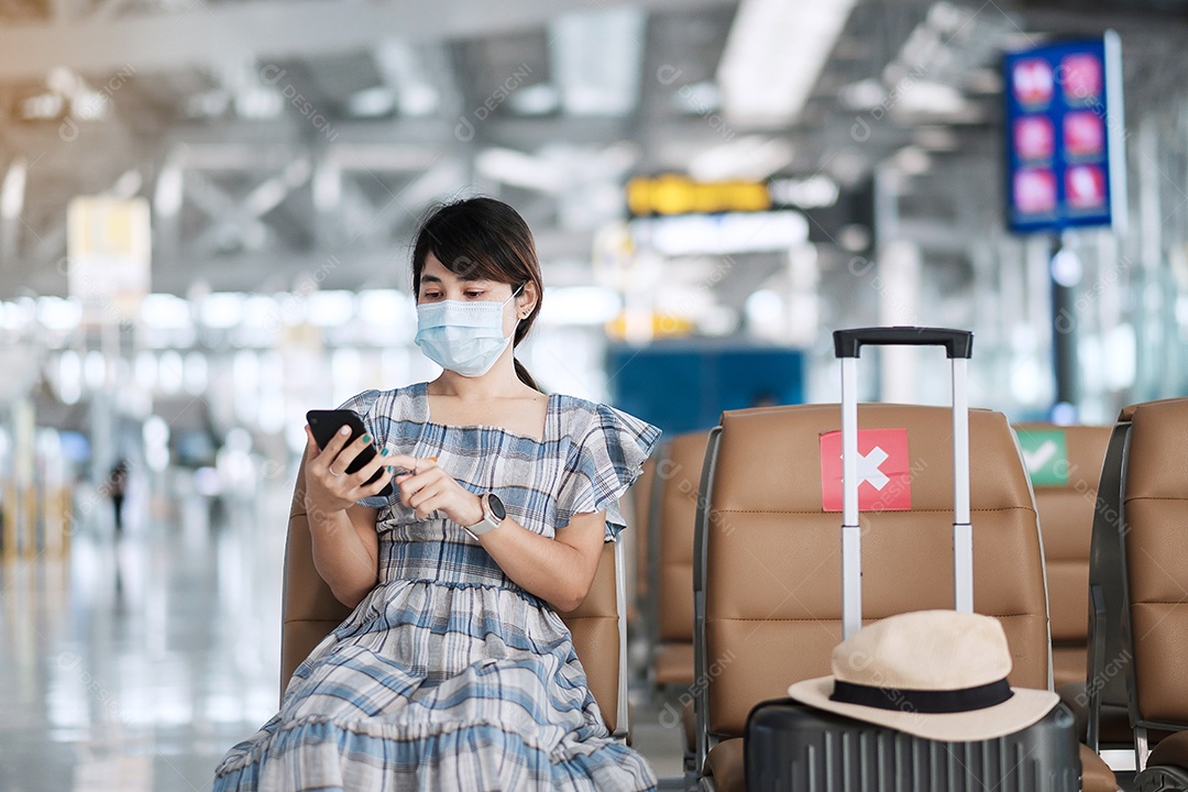 Jovem mulher usando máscara facial e usando smartphone móvel no aeroporto, proteção contra infecção por doença de coronavírus (Covid-19), viajante asiática sentada na cadeira. Novo normal e distanciamento social