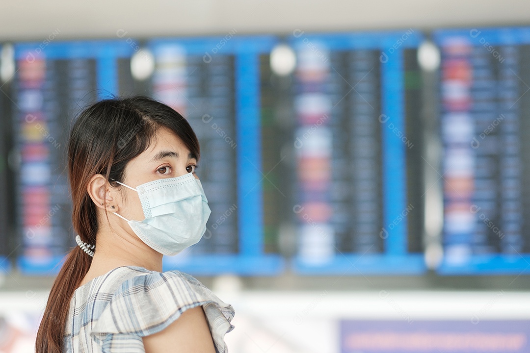 Jovem mulher usando máscara facial cirúrgica verificando o tempo de voo no aeroporto, proteção contra infecção por doença de coronavírus, viajante asiática pronta para viajar. Novo Normal e viagens de segurança sob COVID-19