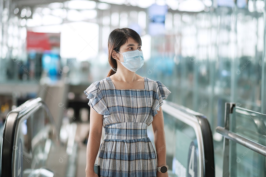 Mulher adulta jovem usando máscara facial no terminal do aeroporto, proteção contra infecção por doença de coronavírus, viajante asiática andando na escada rolante no aeródromo. Novo Normal e viagens sob o conceito COVID-19