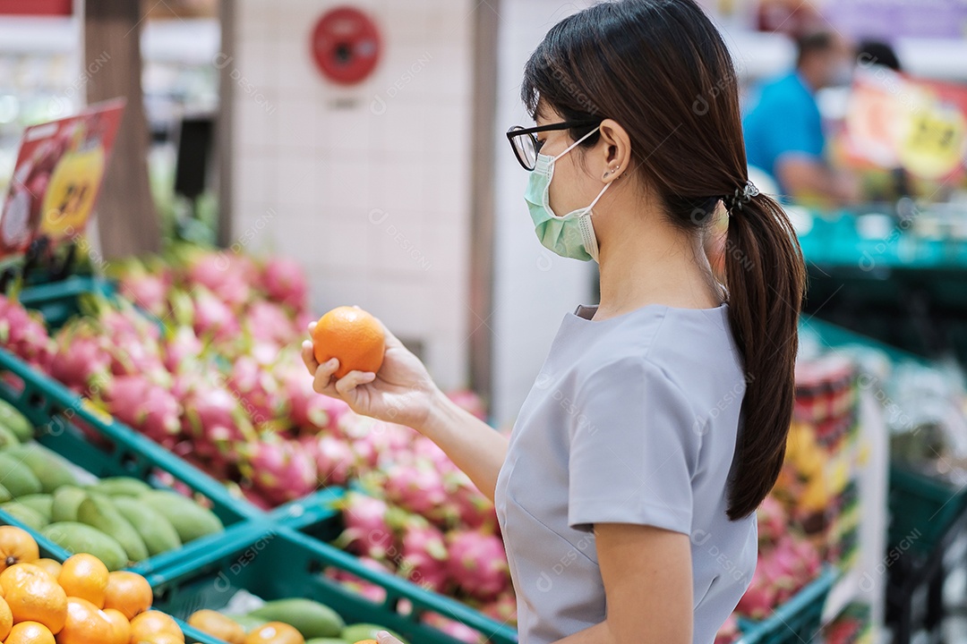 Mulher asiática usando máscara facial cirúrgica e segurando frutas laranja no supermercado ou mercearia, protege a inflexão do coronavírus. Higiene, novo normal e vida sob pandemia de covid-19