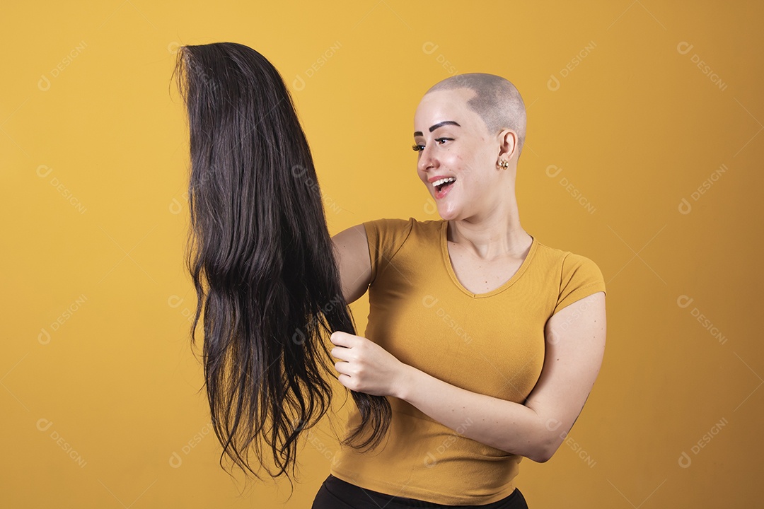 Mulher jovem careca usando blusa bege segurando uma peruca em conceito de câncer, outubro rosa