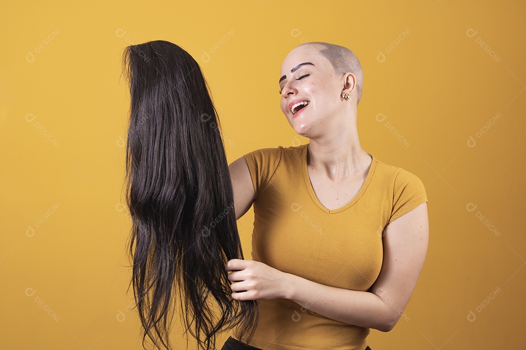 Mulher jovem careca usando blusa bege segurando uma peruca em conceito de câncer, outubro rosa