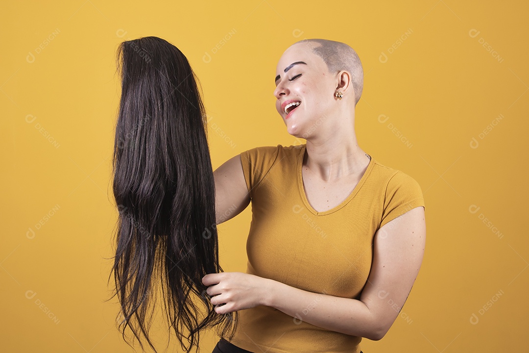 Mulher jovem careca usando blusa bege segurando uma peruca em conceito de câncer, outubro rosa