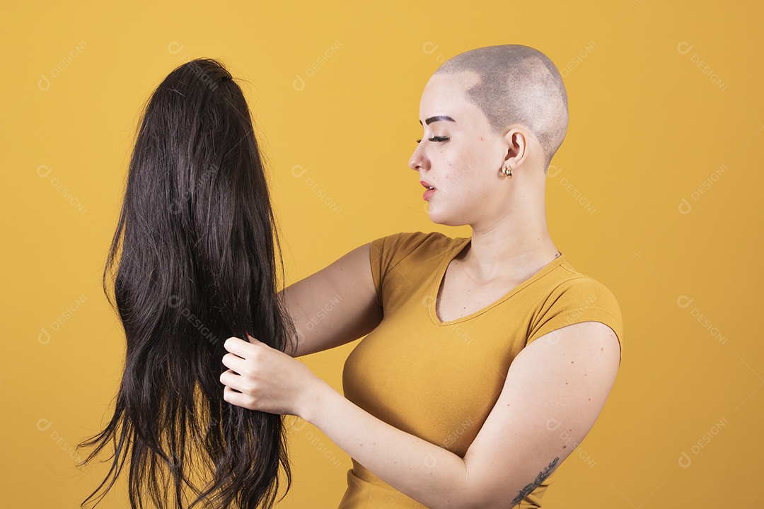 Mulher jovem careca usando blusa bege segurando uma peruca em conceito de câncer, outubro rosa