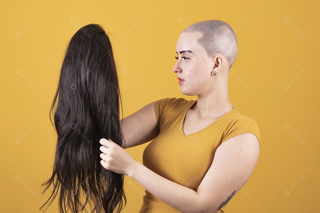 Mulher jovem careca usando blusa bege segurando uma peruca em conceito de câncer, outubro rosa