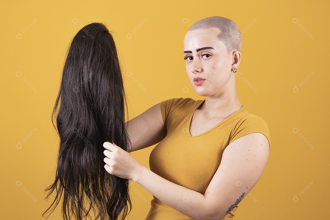 Mulher jovem careca usando blusa bege segurando uma peruca em conceito de câncer, outubro rosa