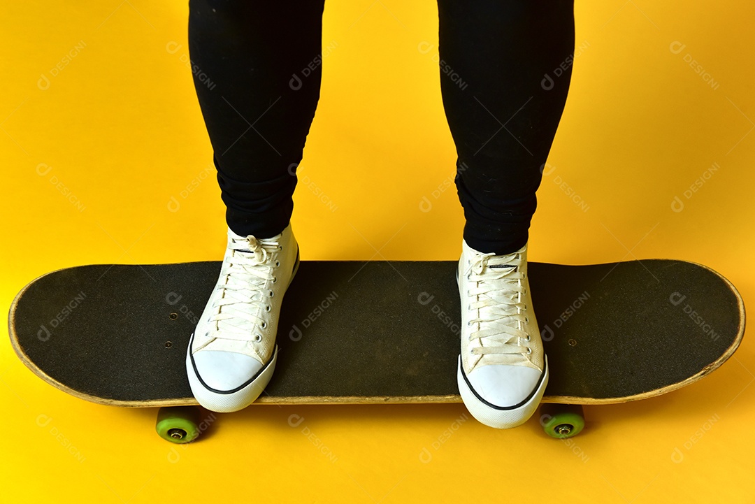 Pernas de garota skatista usando tênis branco sobre fundo amarelo.
