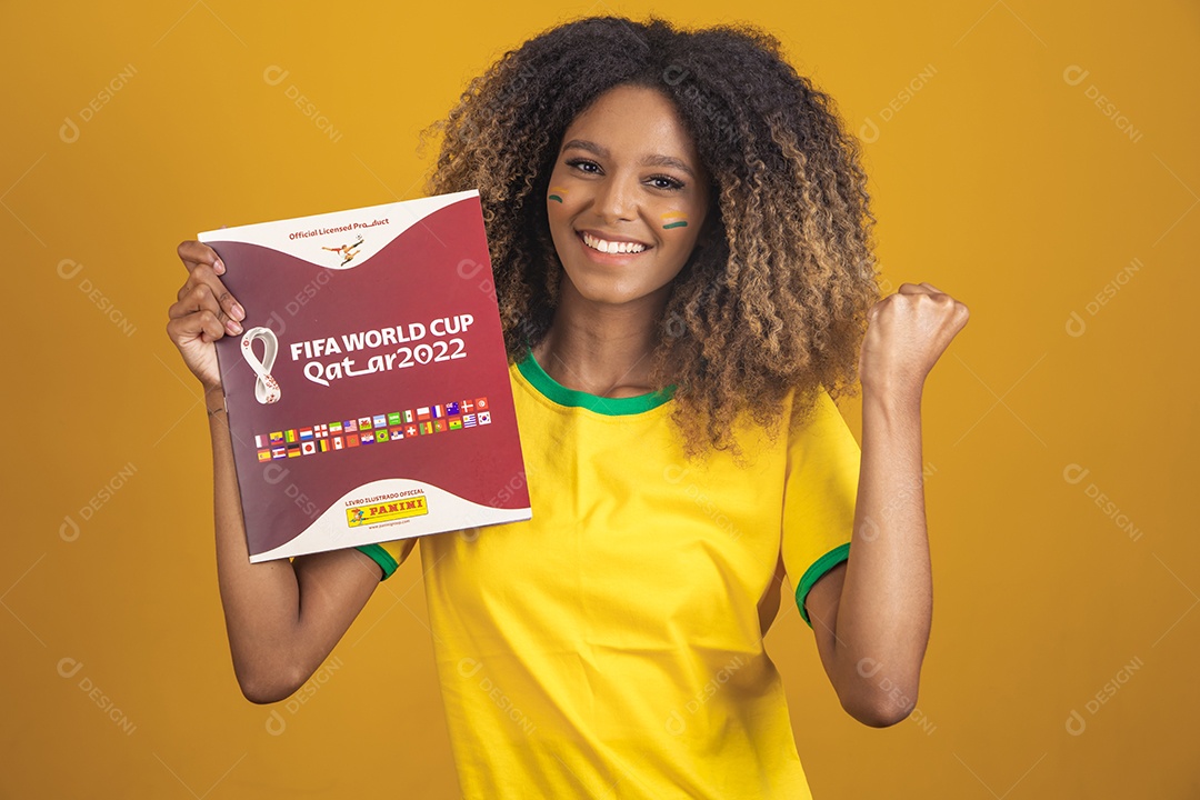 Mulher afro-brasileira vestindo camiseta do Brasil segurando álbum copa do mundo