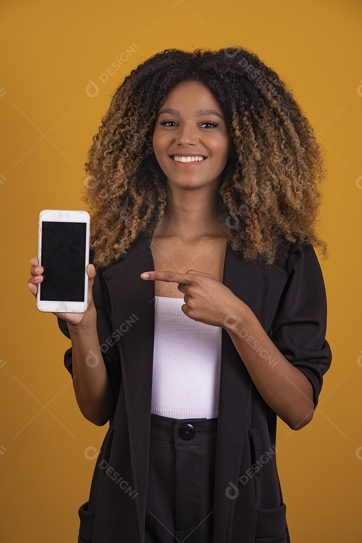 Linda mulher jovem morena cabelo segurando celular smartphone afro sobre fundo isolado amarelo