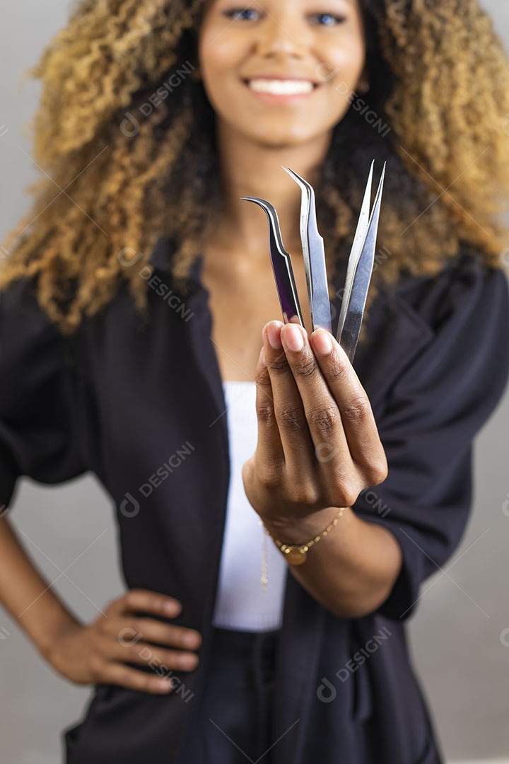 Mulher afro-brasileira vestida formalmente segurando escovinha, pinça e tesoura de sobrancelhas/cílios