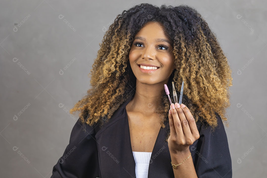 Mulher afro-brasileira vestida formalmente segurando escovinha, pinça e tesoura de sobrancelhas/cílios