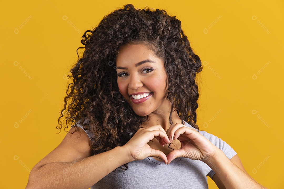 Linda mulher jovem morena cabelo cacheado fazendo gestos coração sobre fundo isolado amarelo
