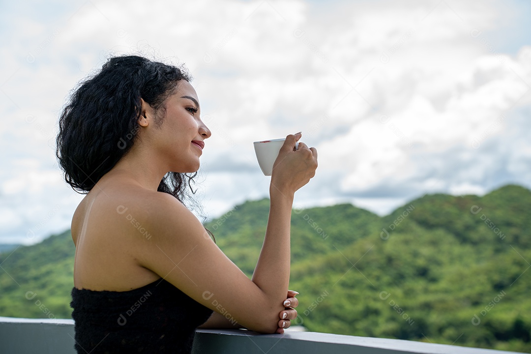 Jovem mulher bebendo uma xícara de café com um sorriso no rosto de pé e sorrindo com um sorriso confiante.