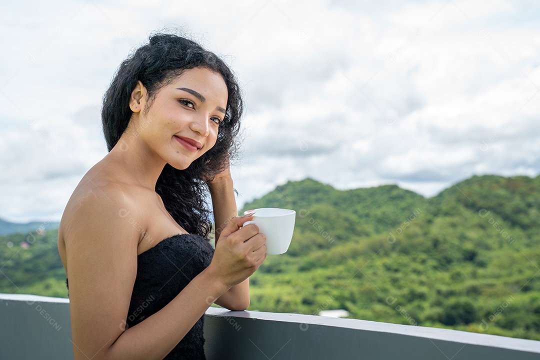 Jovem mulher bebendo uma xícara de café com um sorriso no rosto de pé e sorrindo com um sorriso confiante.