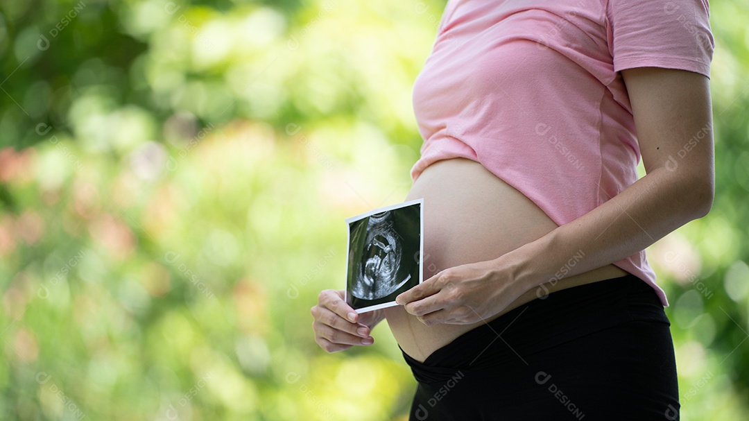 Mulher grávida asiática mostra com bebê de ultrassom, grávida relaxando no parque verde.