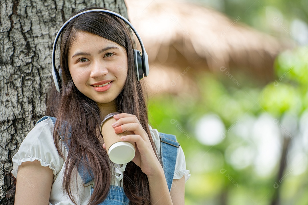 Portrait of girl with headphones and holding coffee cup making gestures in green city park in spri