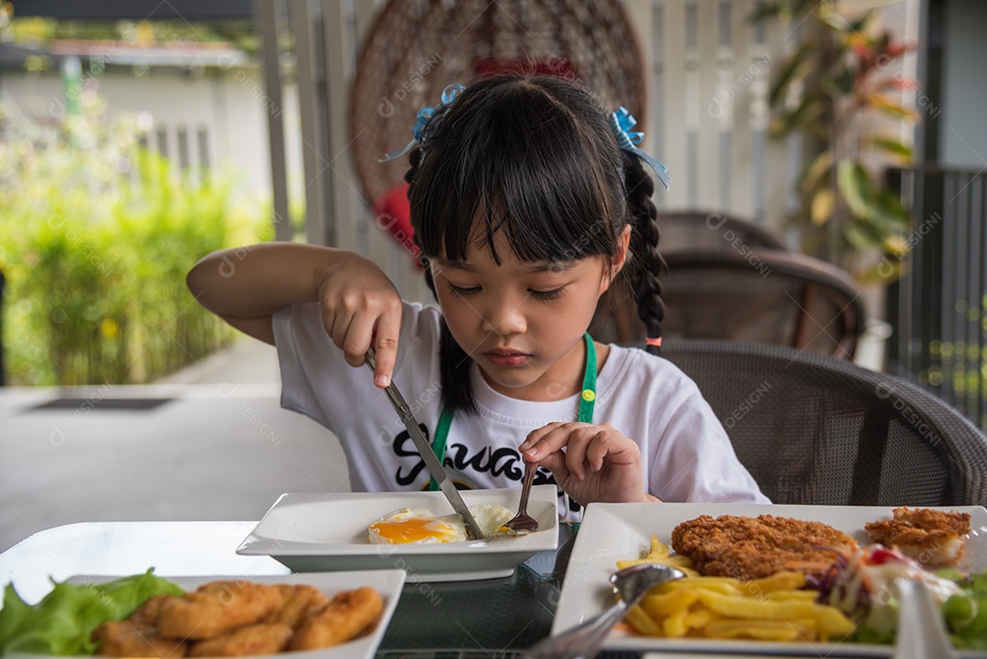 Menina asiática come ovo frito no prato na mesa.