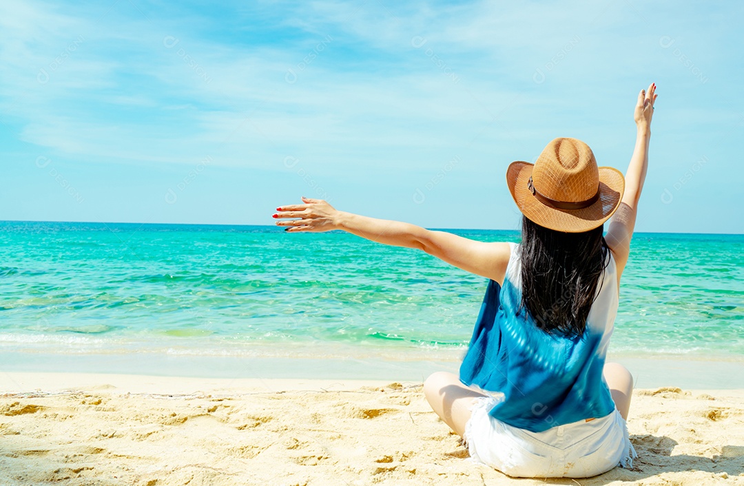 Uma jovem feliz usa chapéu de palha, senta e levanta a mão na praia de areia