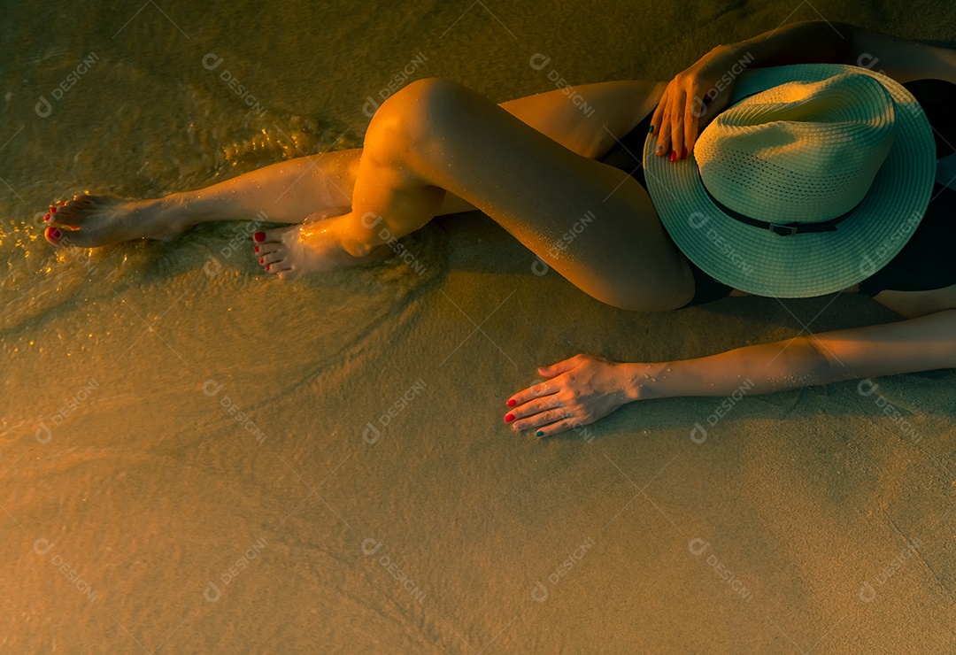 Sexy mulher asiática com chapéu deitado na praia de areia durante o pôr do sol