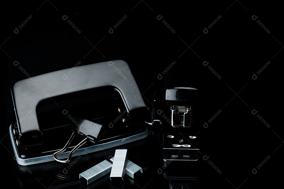 Grampeador, grampos, clipe de papel e perfurador pretos no fundo escuro