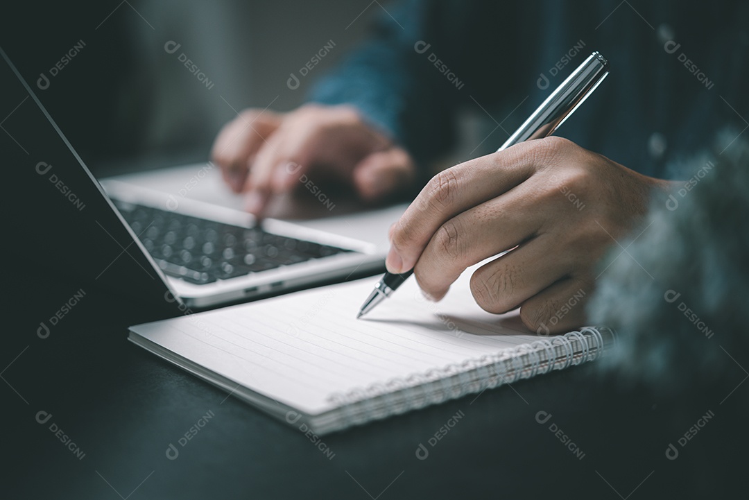 Mão de homem com caneta escrevendo no notebook com computador portátil na mesa.