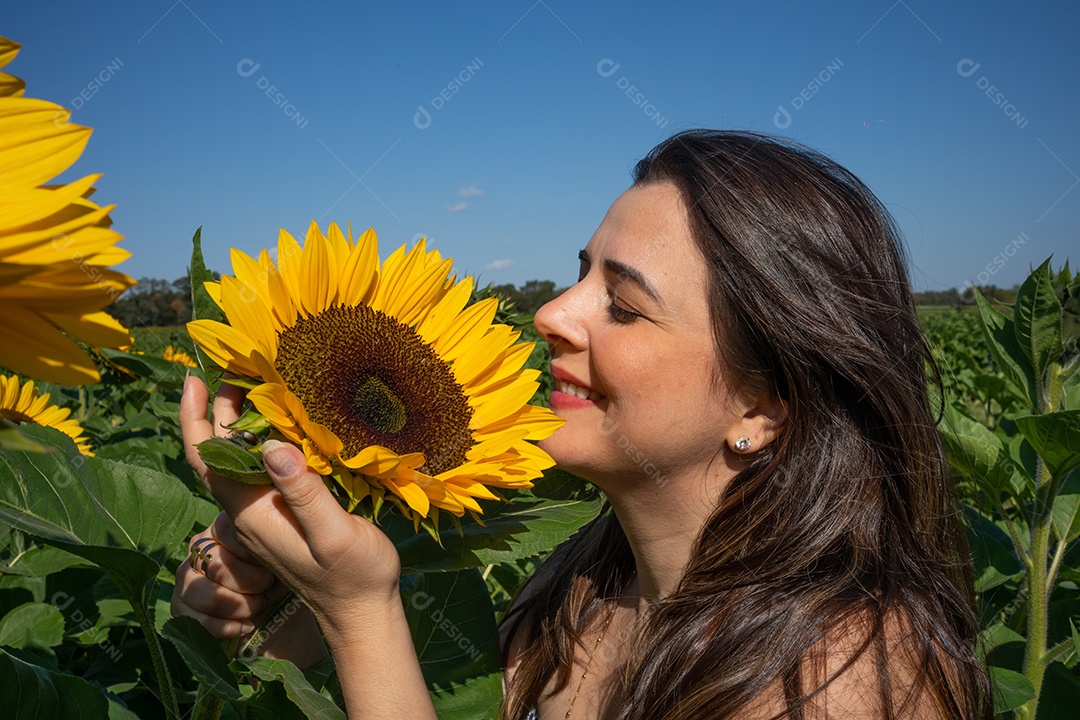 Mulher sorrindo e cheirando girassol em uma plantação.